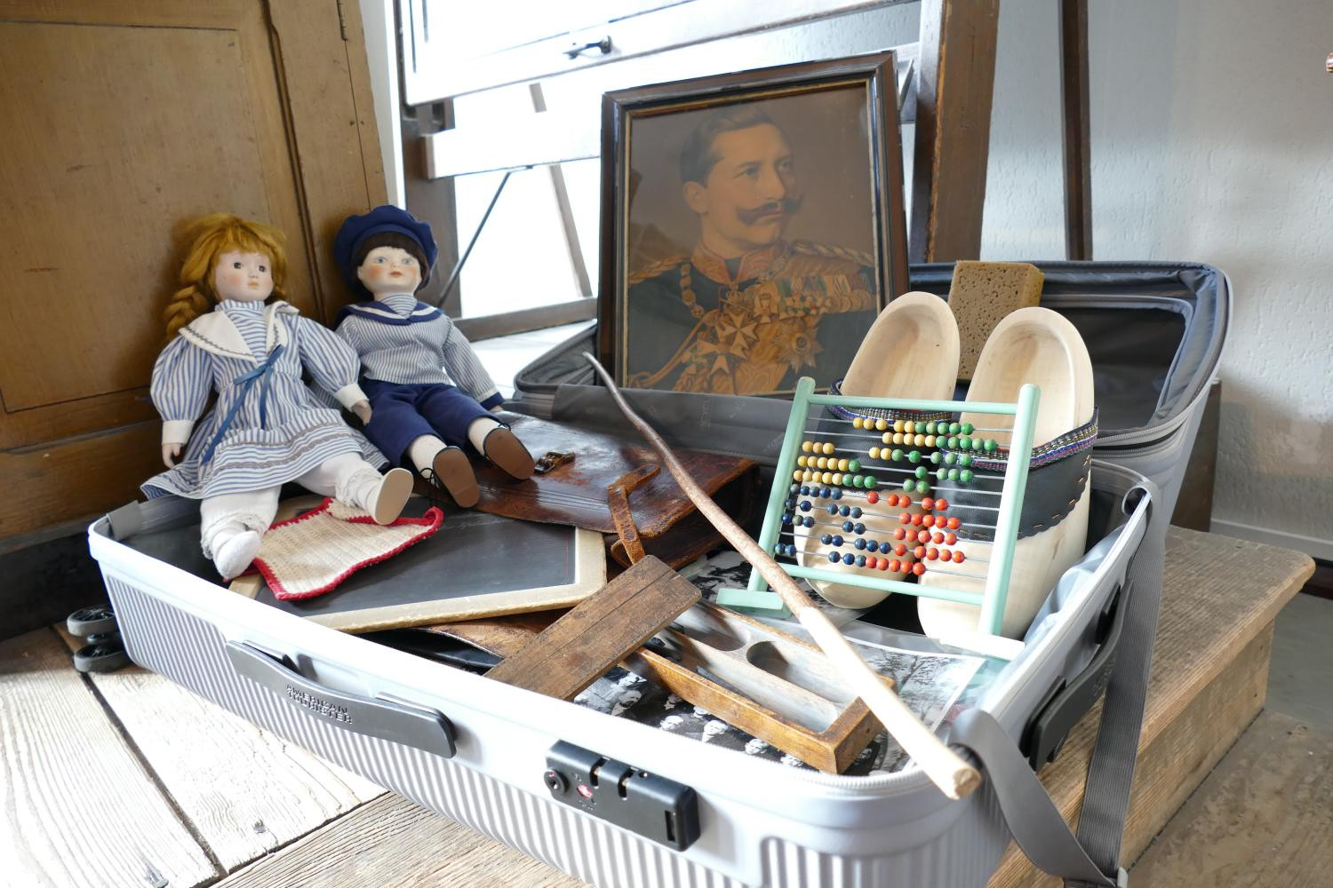 Das Foto eines geöffneten Koffers, in dem sich Holzschuhe, zwei Puppen, ein Portraitbild von Kaiser Wilhelm II, ein Schwamm, ein Rechenschieber eine Kreidetafel und Schwarz-weiß Fotografien befinden.