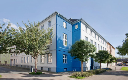 Ein Eckhaus, fotografiert von dem Platz, der vor dem Haus liegt, aus. Das Haus ist dreigeschossig mit einer weißen Fassade. Die Ecke des Hauses ist blau gestrichen. Vor dem Haus stehen mehrere Straßenbäume.