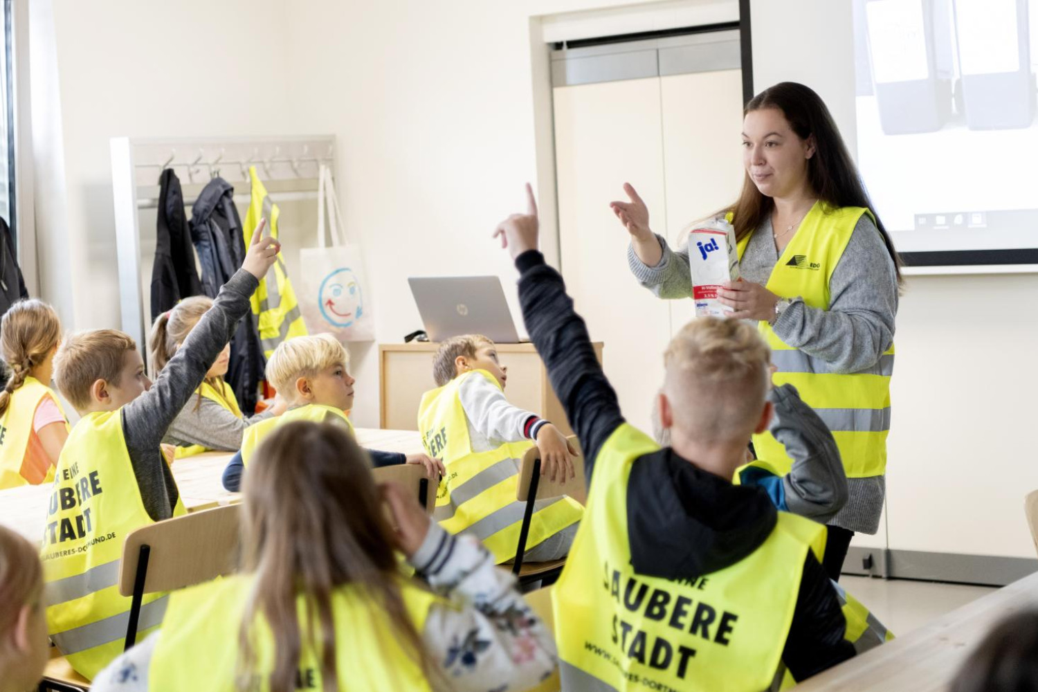 Eine Gruppe Schüler*innen sitzt an Konferenztischen. Alle Kinder haben eine gelbe Warnweste mit einem Schriftzug auf dem Rücken an. Die Worte "saubere Stadt" sind groß geschrieben und daher zu lesen. Vor den Kindern steht eine Frau, die ebenfalls eine Warnweste trägt. In der Hand hält sie ein Tetrapack. Zwei Kinder zeigen auf.