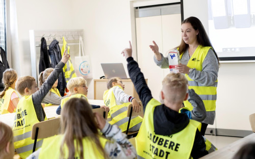 Eine Gruppe Schüler*innen sitzt an Konferenztischen. Alle Kinder haben eine gelbe Warnweste mit einem Schriftzug auf dem Rücken an. Die Worte "saubere Stadt" sind groß geschrieben und daher zu lesen. Vor den Kindern steht eine Frau, die ebenfalls eine Warnweste trägt. In der Hand hält sie ein Tetrapack. Zwei Kinder zeigen auf.