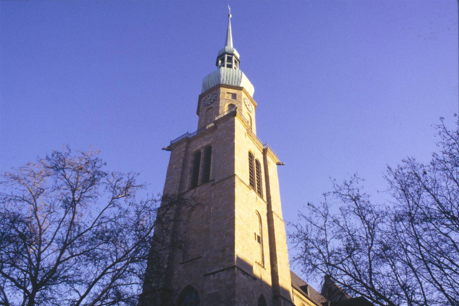 Der Turm der Reinoldikirche in der Ansicht von unten. Um den Turm herum stehen Bäume, die kein Laub tragen. Der Himmel ist blau und die rechte Seite des Turms wird von der Sonne beschienen.