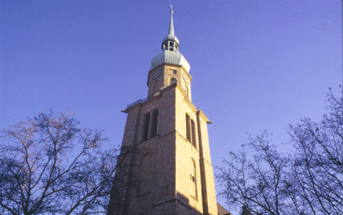 Der Turm der Reinoldikirche in der Ansicht von unten. Um den Turm herum stehen Bäume, die kein Laub tragen. Der Himmel ist blau und die rechte Seite des Turms wird von der Sonne beschienen.