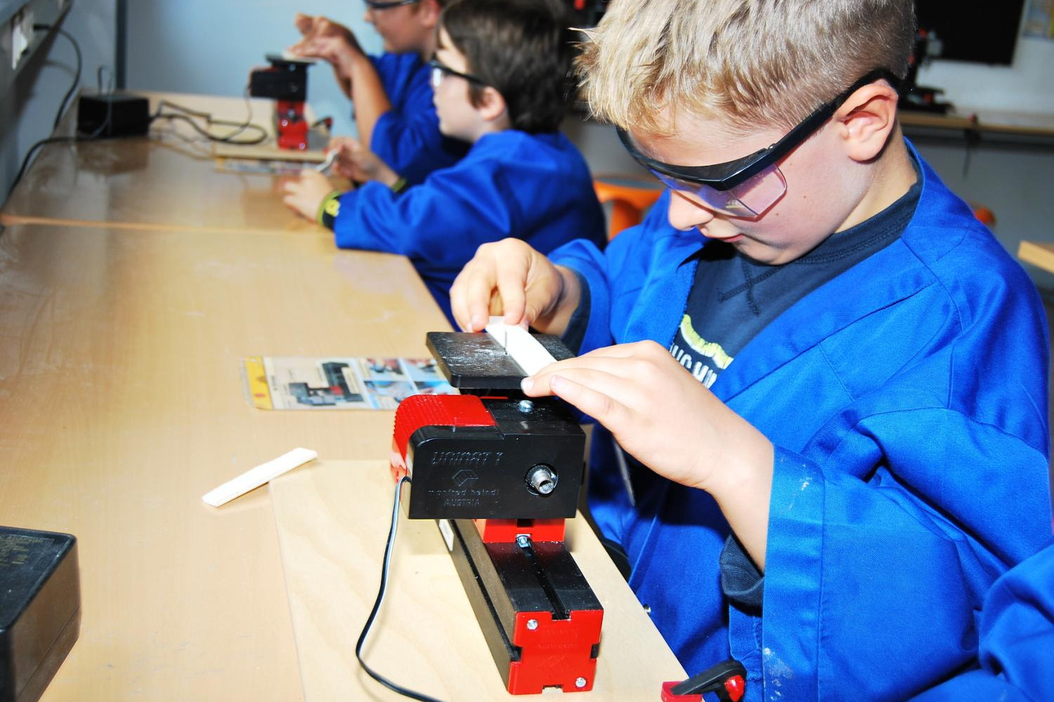 Mehrere Kinder stehen an einer Werkbank. Alle tragen blaue Kittel und eine Schutzbrille. Auf der Werkbank sind kleine Stichsägen befestigt. Die Kinder bearbeiten damit Holz.