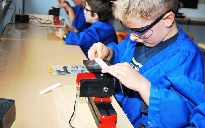 Mehrere Kinder stehen an einer Werkbank. Alle tragen blaue Kittel und eine Schutzbrille. Auf der Werkbank sind kleine Stichsägen befestigt. Die Kinder bearbeiten damit Holz.