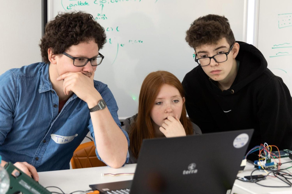 Drei Jugendliche schauen gemeinsam auf den Bildschirm eines Laptops. Zwei von ihnen sind in einer Denkerpose mit einer Hand vor dem Kinn. Hinter den Personen ist ein Whiteboard mit grüner Schrift. Neben dem Laptop einige Kabel.