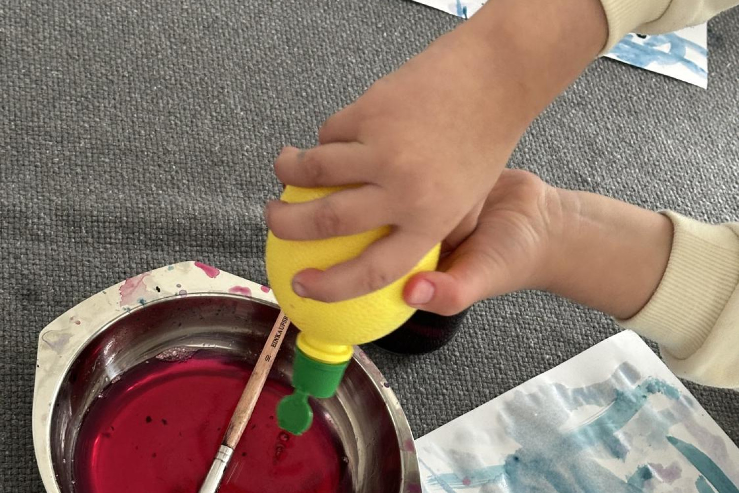 Das Bild zeigt Kinderhände, die Farben anmischen. Ein Kind hält Zitronensaft in der typischen Verpackung in den Händen und drück diesen in den Farbtopf.