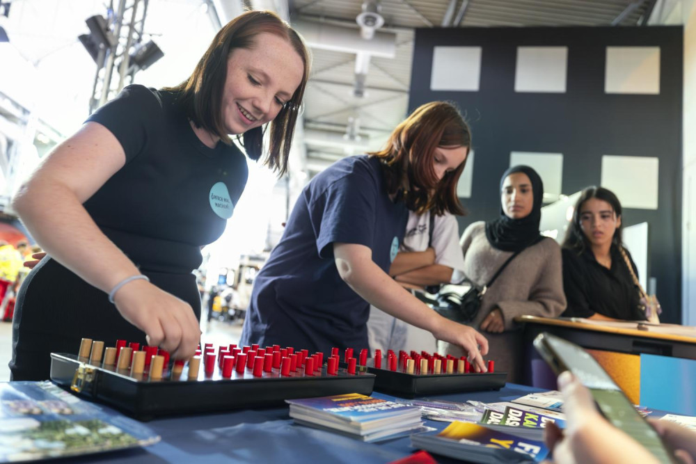 Zwei Jugendliche stehen vor einem Spiel und spielen dieses, zwei weiter Jugendliche sehen dabei zu.