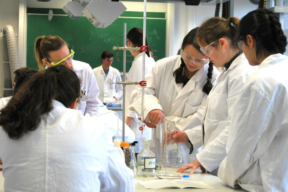 Eine Gruppe Mädchen steht, mit Laborkitteln und Schutzbrillen bekleidet vor einem Versuchsaufbau mit mehreren Gefäßen. Eines der Mädchen schüttet etwas von einem Gefäß in ein anderes.