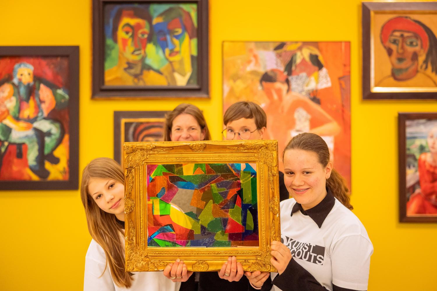 Vier Personen - zwei Erwachsene und zwei Jugendliche - halten ein buntes, Bild mit vielen unterschiedlichen Formen hoch. Im Hintergrund ist eine strahlend gelbe Wand, an der verschiedene impressionistische Kunstwerke hängen.