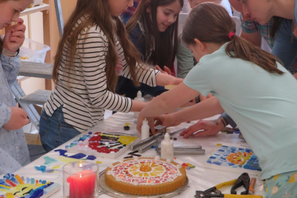 Eine Gruppe Mädchen steht gemeinsam mit einer Frau um einen Tisch herum. Jedes Kind gestaltet ein Mosaik aus Glassteinen. In der Mitte des Tisches steht ein Kuchen.