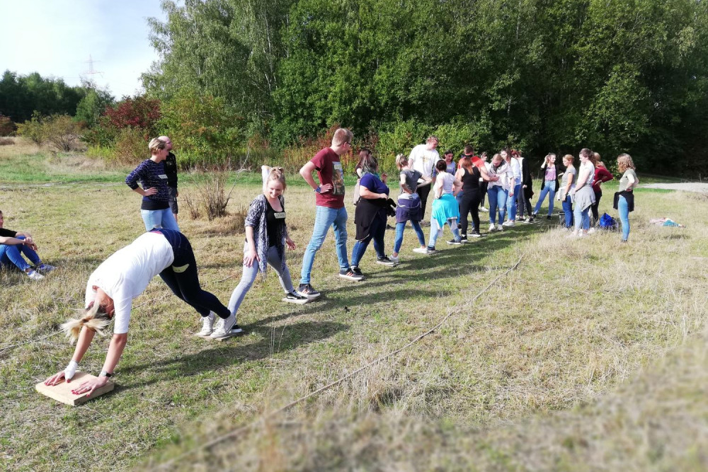 Eine Gruppe Jugendlicher steht auf einer Wiese, jeder der Jugendlichen steht mit einem Fuß auf einem Holzbrett und mit dem anderen Fuß auf einem anderen Brett. Dadurch verbinden die Jugendlichen alle Bretter miteinander. Im Hintergrund stehen zwei Erwachsen