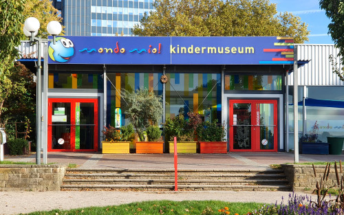 Die Fassade des mondomio Kindermuseums. DAs Gebäude ist ein Pavillion mit eineer Glasfront. Die Rahmen der Türen sind leuchtend rot und der Schriftzug mondomio bunt. Vor dem Pavillion ist eine Terasse, die über vier Stufen zu erreichen ist. Im Hintergrund sind zwei Bäume und ein Hochhaus