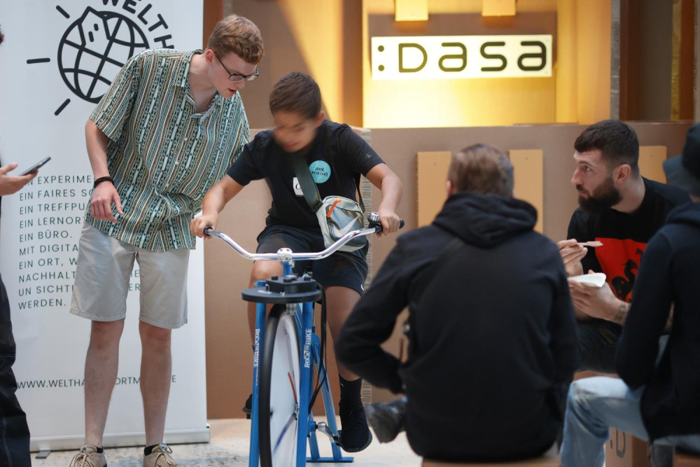 Ein Kind sitzt auf einem Fahrrad, das allerdings festgestellt ist und nur vorn ein Rad hat. Das Rad steht in einer Ausstellung. Neben dem Jungen steht ein anderer Junge und sagt etwas zu ihm. Ein Mann, der daneben sitzt und etwas isst schaut die beiden an.