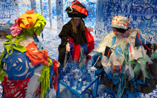 Drei Jugendliche sind mit aufwendigen, bunten Kostümen verkleidet und stehen in einem blau-weißen Szenenbild. Sie stehen um einen kleinen Beistelltisch herum, auf dem viele blaue Farbflaschen stehen. In diese tauchen sie Pinsel.