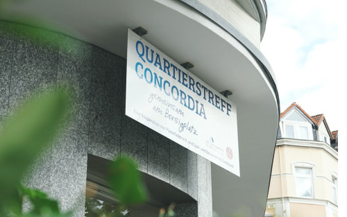 Ein Bildausschnitt mit einem Teil einer grauen Fassade. Über einem Fenster hängt ein Schild mit der Aufschrift: Quartierstreff Concordia gemeinsam am Borsigplatz.