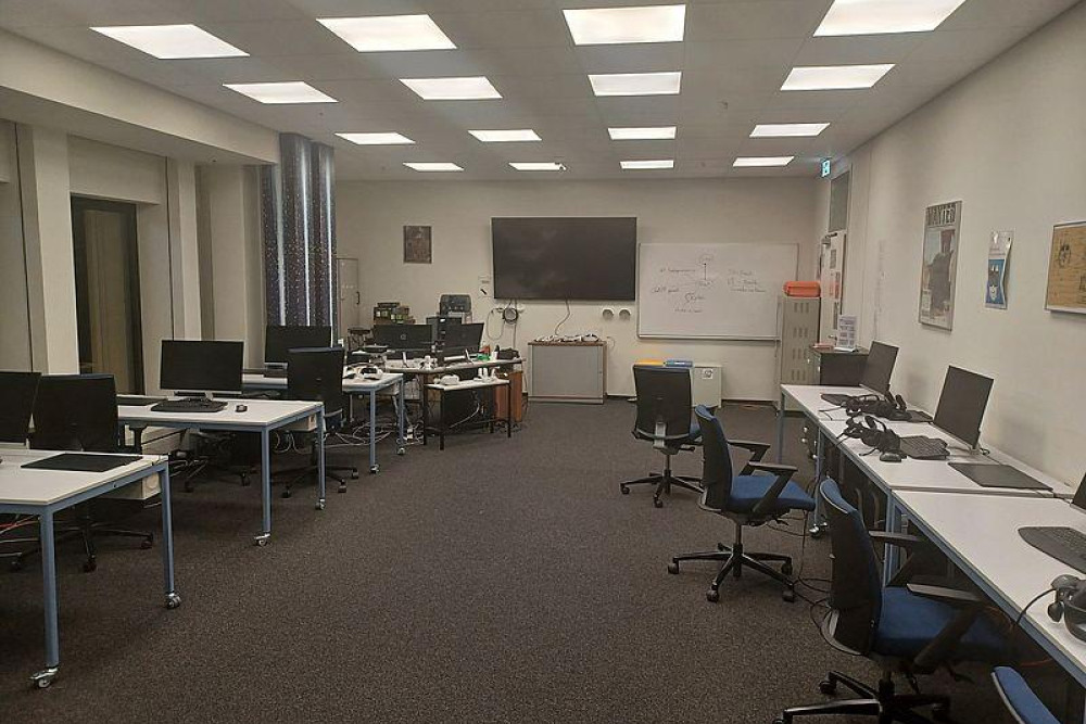 Ein Raum mit Tischen, an denen Arbeitsplätze mit Bildschirmen eingerichtet sind. An der Wand hängen eine Tafel und ein Whiteboard.