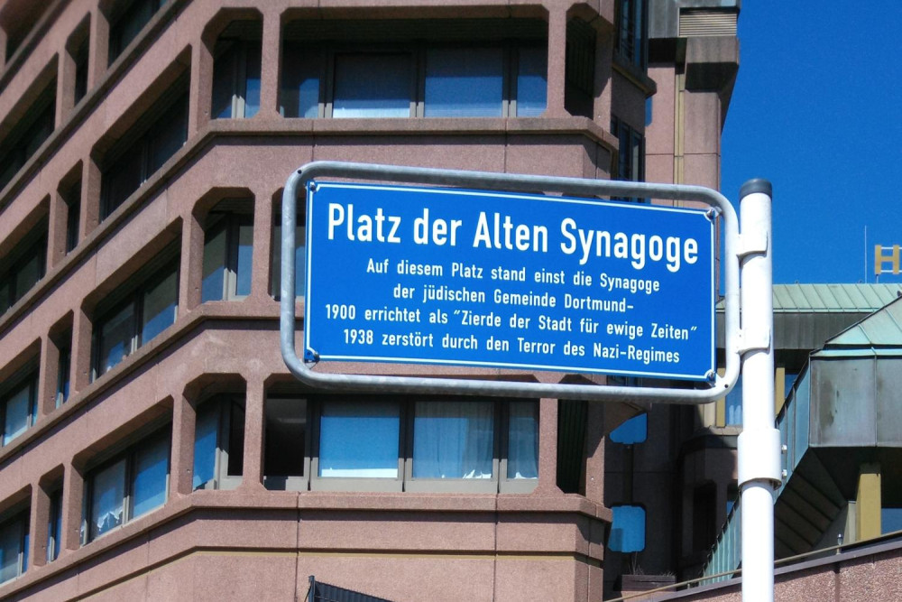 Ein Straßenschild mit dem Text: Platz der alten Synagoge - Auf diesem Platz stand einst eine Synagoge der jüdischen Gemeinde Dortmund - 1900 errichtet als Zierde der Stadt für ewige Zeiten 1938 zerstört durch den Terror des Nazi-Regimes
