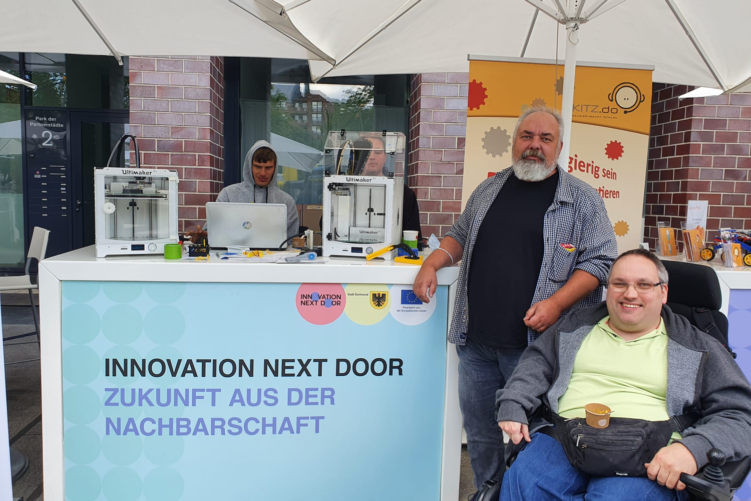 Ein Messestand ist vor der Fassade eines verklinkerten Gebäudes aufgebaut. Auf dem Tresen stehen zwei 3D-Drucker und ein Laptop. Hinter dem Tresen stehen zwei Männer und schauen auf den Laptop und den Drucker. Vor dem Tresen hängt ein Banner. Auf diesem ist der Schriftzug: Innovation next door - Zukunft aus der Nachbarschaft. Neben dem Tresen steht ein Mann. ein zweiter Mann sitzt neben ihm in einem Rollstuhl. Beide sehen in die Kamera.