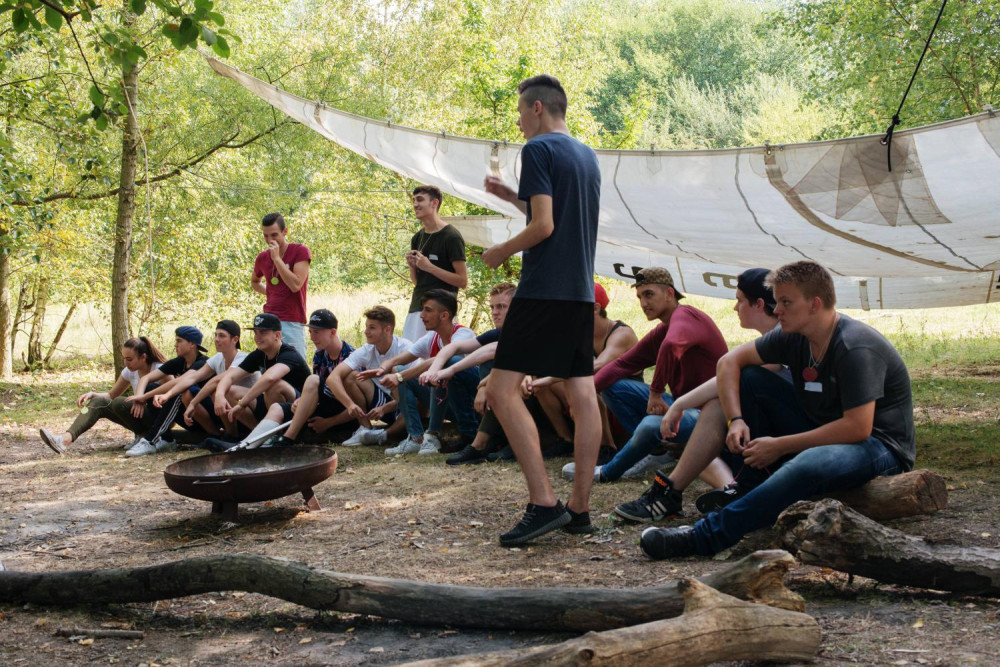 Eine Gruppe Jugendlicher sitzt auf entrindeten Ästen, die um eine Feuerschale herum platziert sind. Hinter ihnen ist ein Sonnensegel zwischen zwei Bäumen aufgespannt.