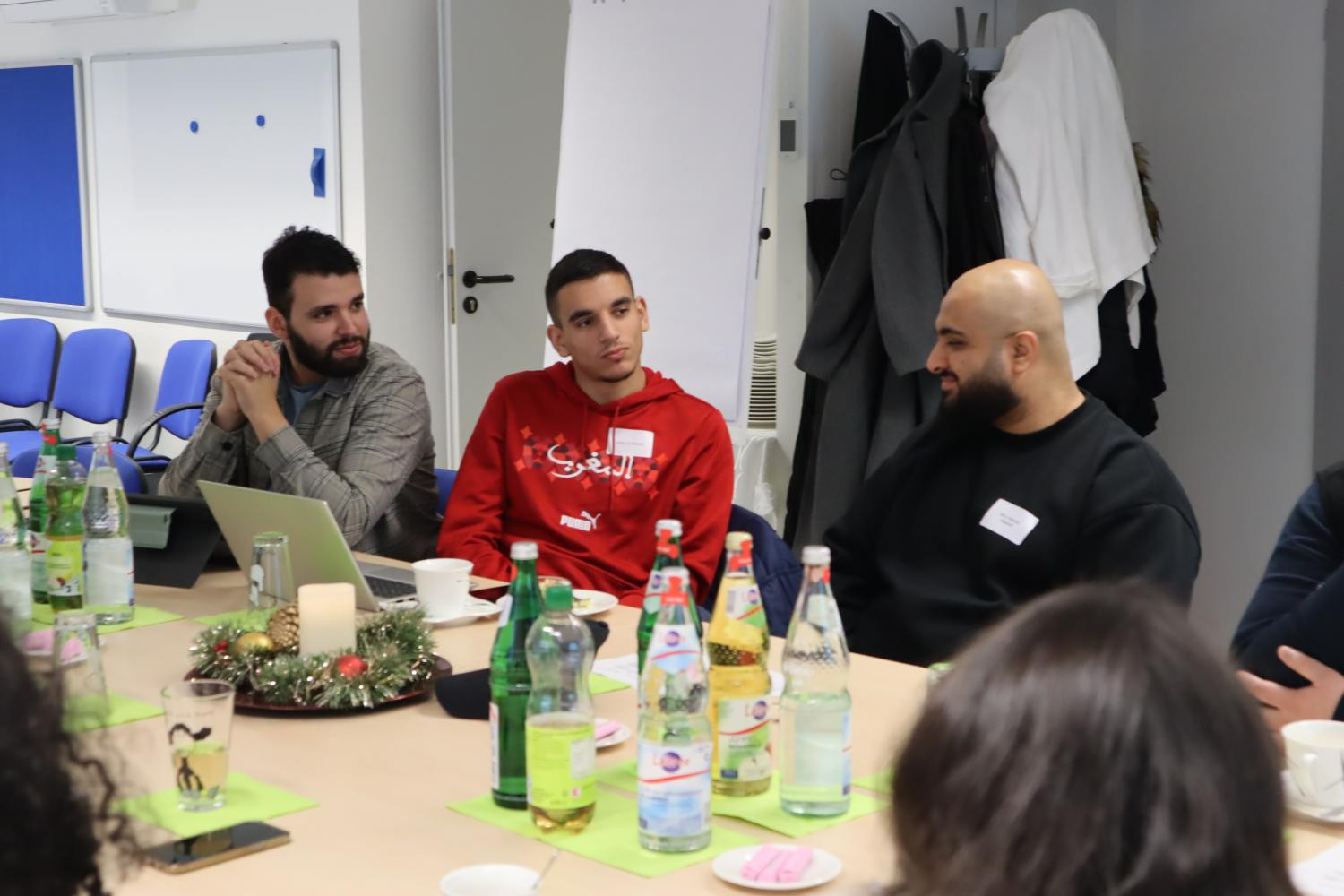 Drei junge Männer sitzen nebeneinander an einem Konferenztisch. Zwei von ihnen schauen den dritten an. An dem Tisch sitzen noch drei weitere Personen, von denen allerdings nur die Haare oder ein Arm zu sehen sind.