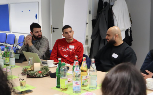 Drei junge Männer sitzen nebeneinander an einem Konferenztisch. Zwei von ihnen schauen den dritten an. An dem Tisch sitzen noch drei weitere Personen, von denen allerdings nur die Haare oder ein Arm zu sehen sind.