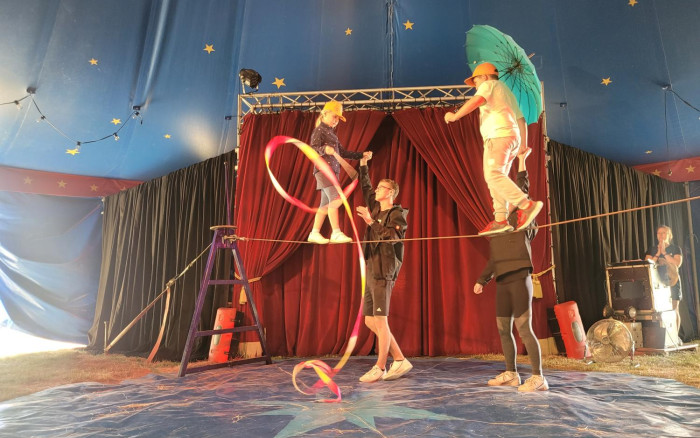 In einer Zirkusmanege ist ein Seil auf etwa einem Meter Hohe gespannt. Zwei Kinder balancieren über das Seil. ein Kind hat ein Gymnastikband in der Hand, das andere Kind eine großen Schirm. Neben den Kindern steht jeweils ein Erwachsener und gibt Hilfestellungen.