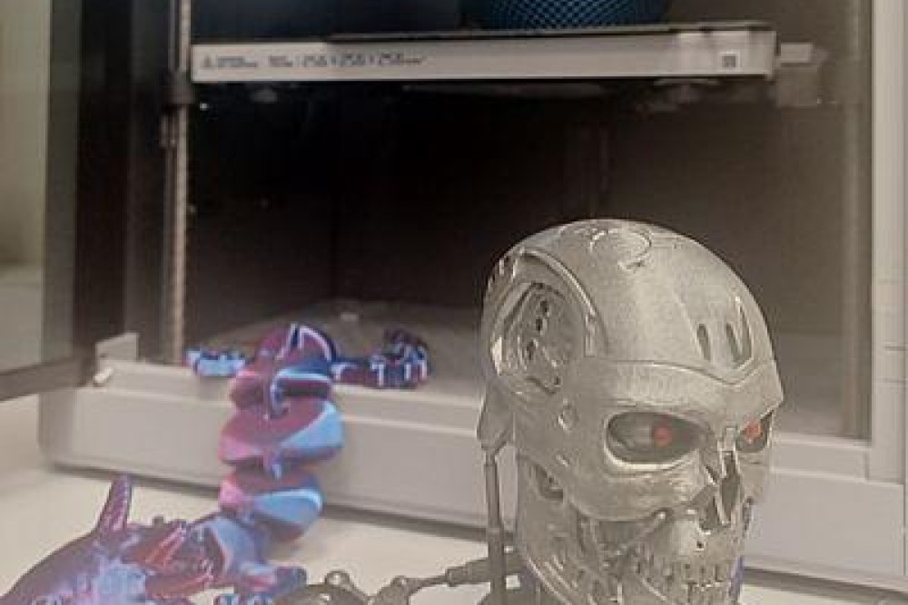 Ein 3D-Drucker mit zwei Druckerzeugnissen: ein Fischkopf an einer langen Kette und ein Stahlschädel mit roten Augen.